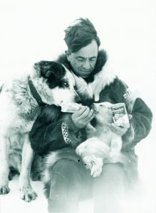 A man wearing warm clothing feeding a baby sea with a dog beside him.