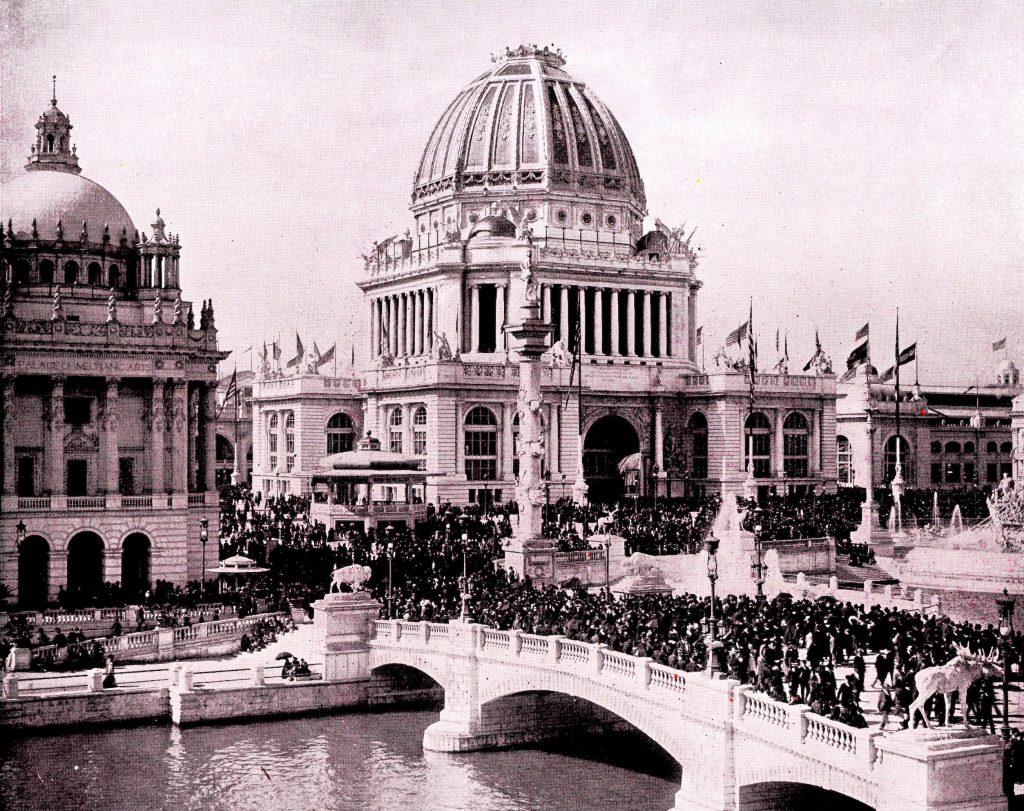 A photo of the administration building, a building with a large domed top surrounded by numerous flags. There are many fairgoers around the building and on a bridge that goes over a river in the foreground.