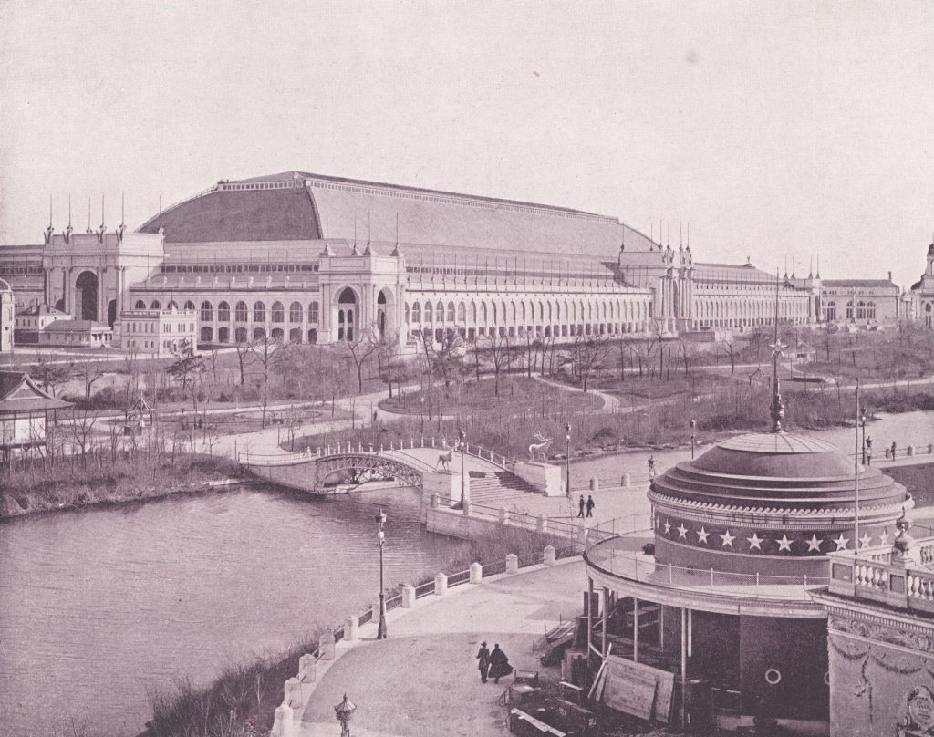 A large building (the Manufactures building) with arched doorways around the perimeter lies in the background with a river crossing a bridge in the foreground