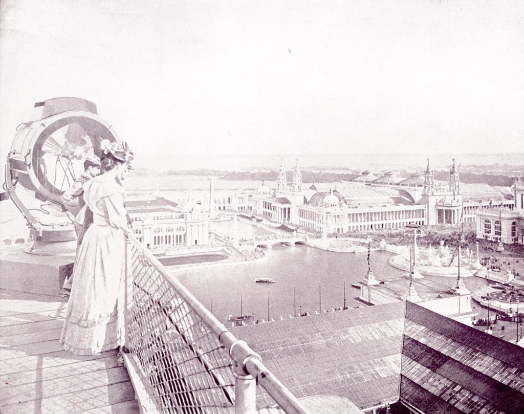 two fairgoers stand at the fence on the top of the Manufactures building, look down on a dock with boats as well as some buildings of the fair