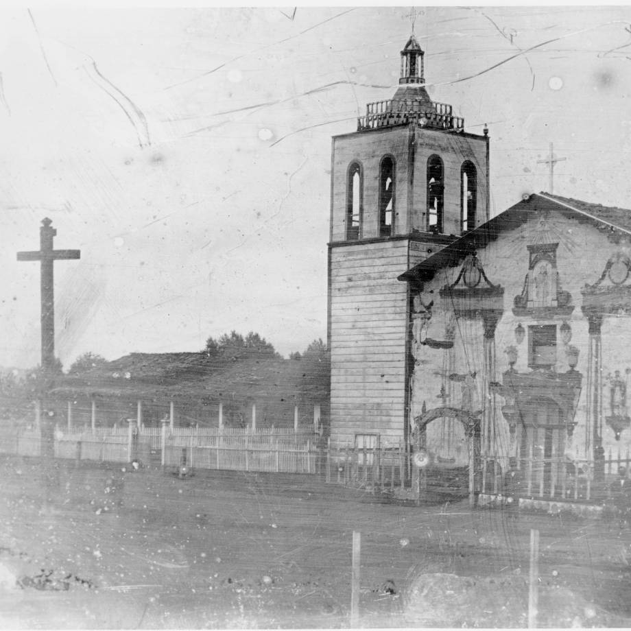 A black and white image of the fifth and final adobe Santa Clara Mission church. On the left side of the image is a large wooden cross, while on the right is the mission itself, constructed mostly of adobe, but the bell tower is encompassed by wood. Credit:  SCU Digital Collection.