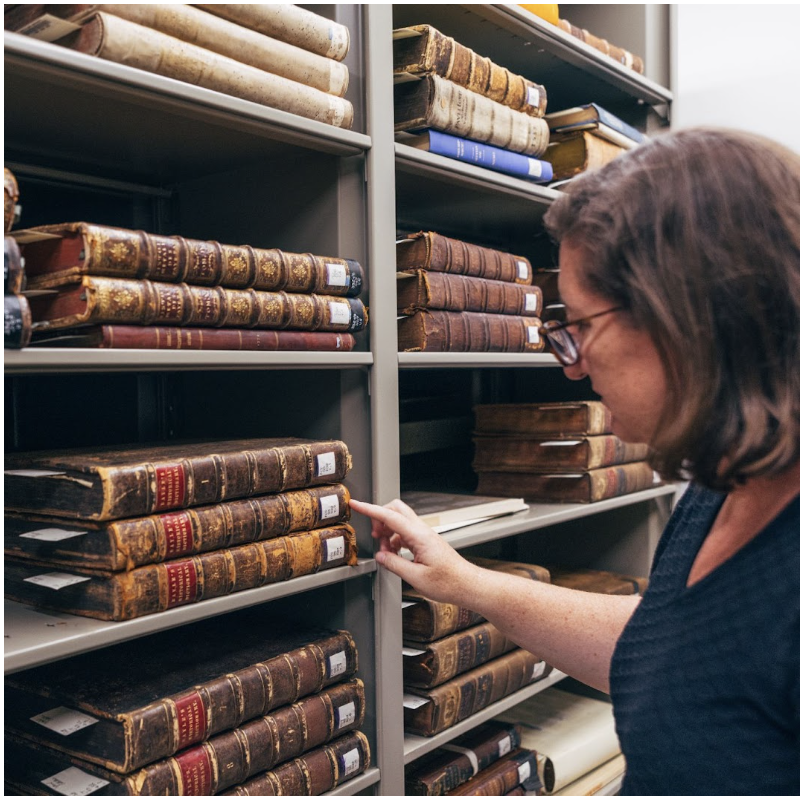 On the left side of the photo is a shelf housing various texts in the Santa Clara University Archive, with Monica Keane, the Archives & Special Collections Librarian, looking through them on the left. Credit: Photo by Miguel Ozuna. 