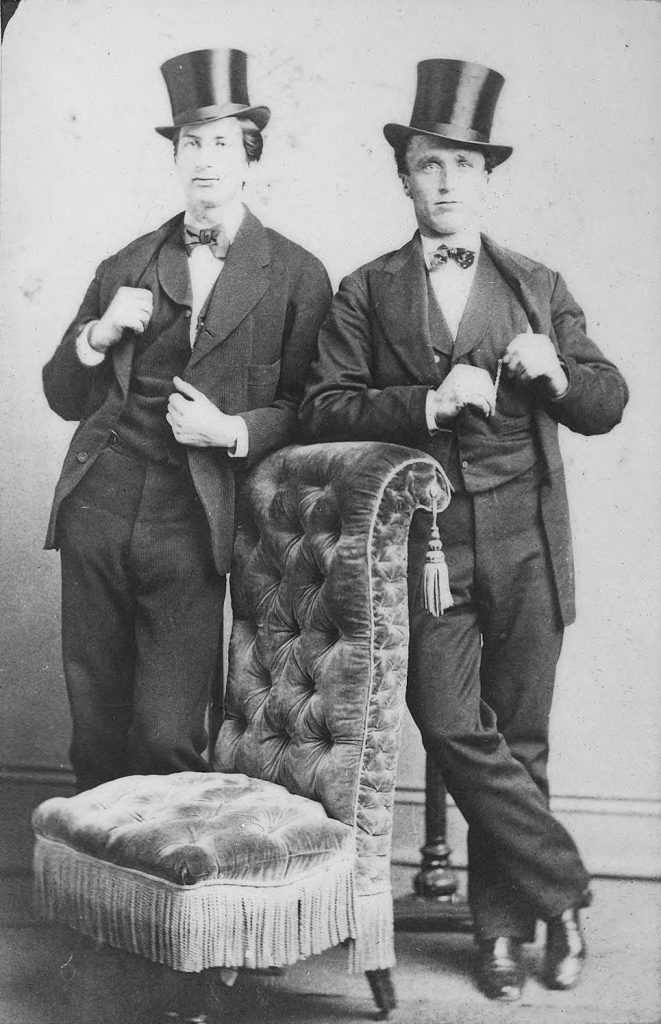 Two young men in top hats posing next to a velvet chair