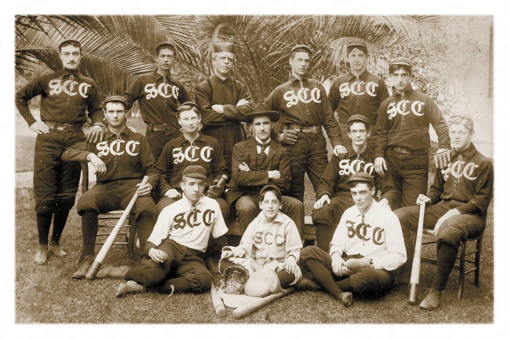 Boys sitting on the ground in baseball uniforms around their coach in a suit