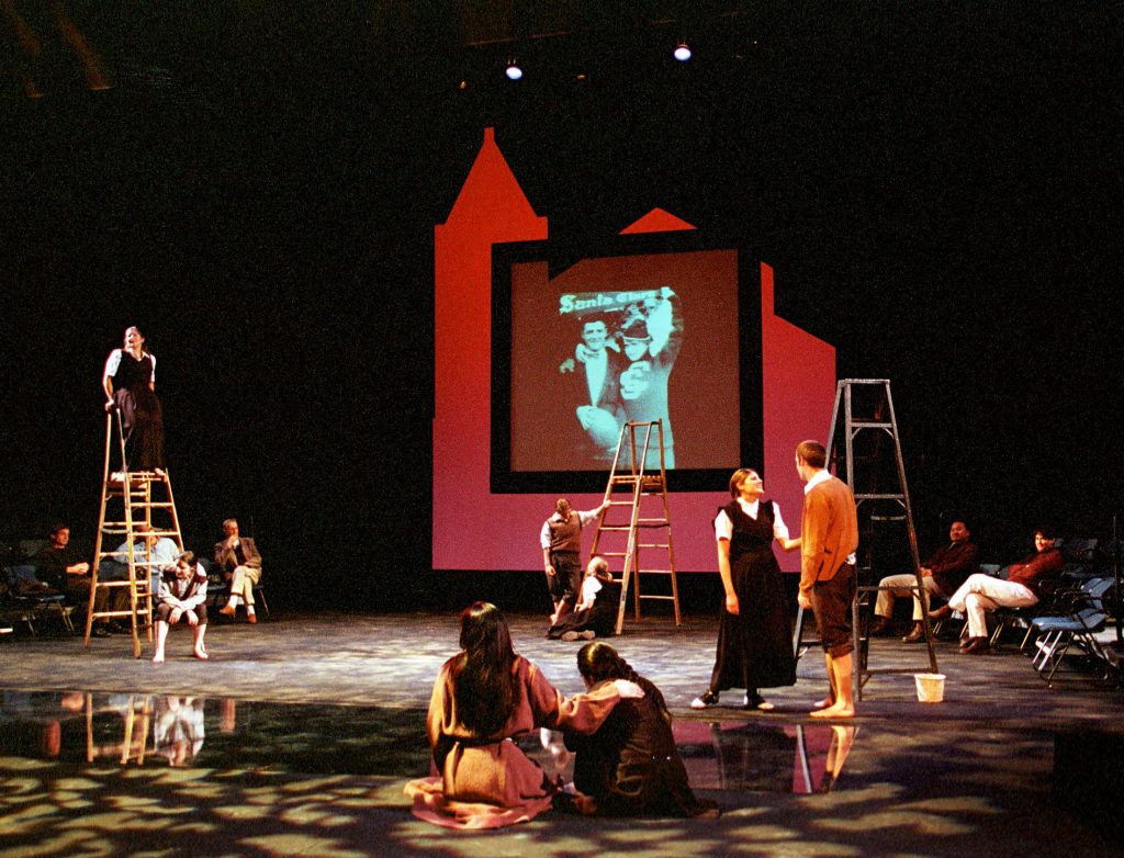 Full stage with actors talking, ladders and a large red screen in the background.