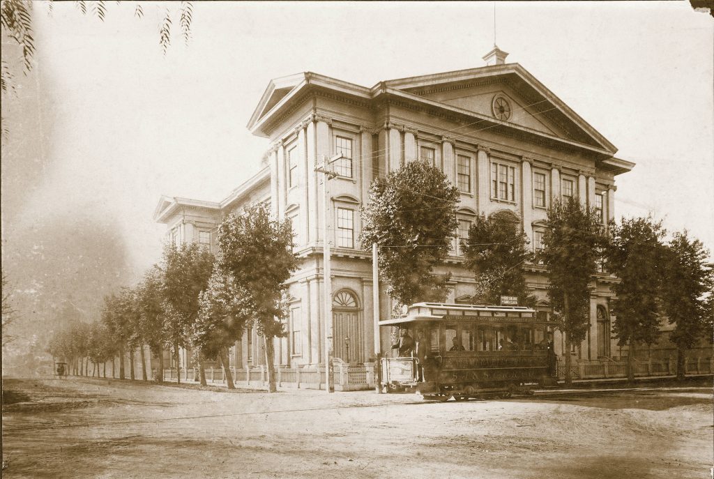 Italianate Victorian building with trees and a trolley in front.